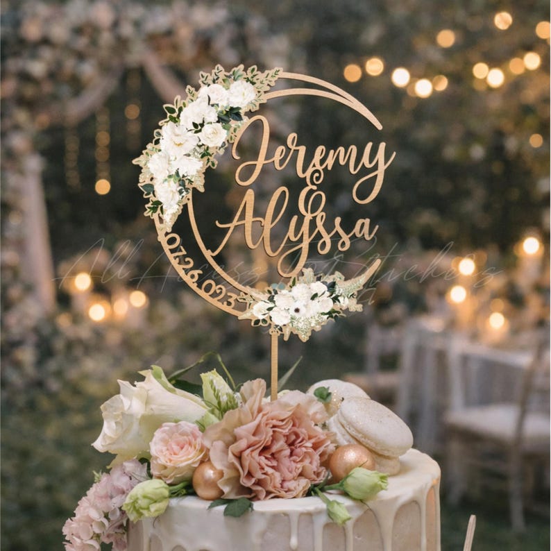 May include: A circular cake topper featuring the names Jeremy and Alyssa in a gold script font, with the date 07.26.023 engraved along the side, decorated with white floral arrangements on the top left and bottom right sections of the ring.