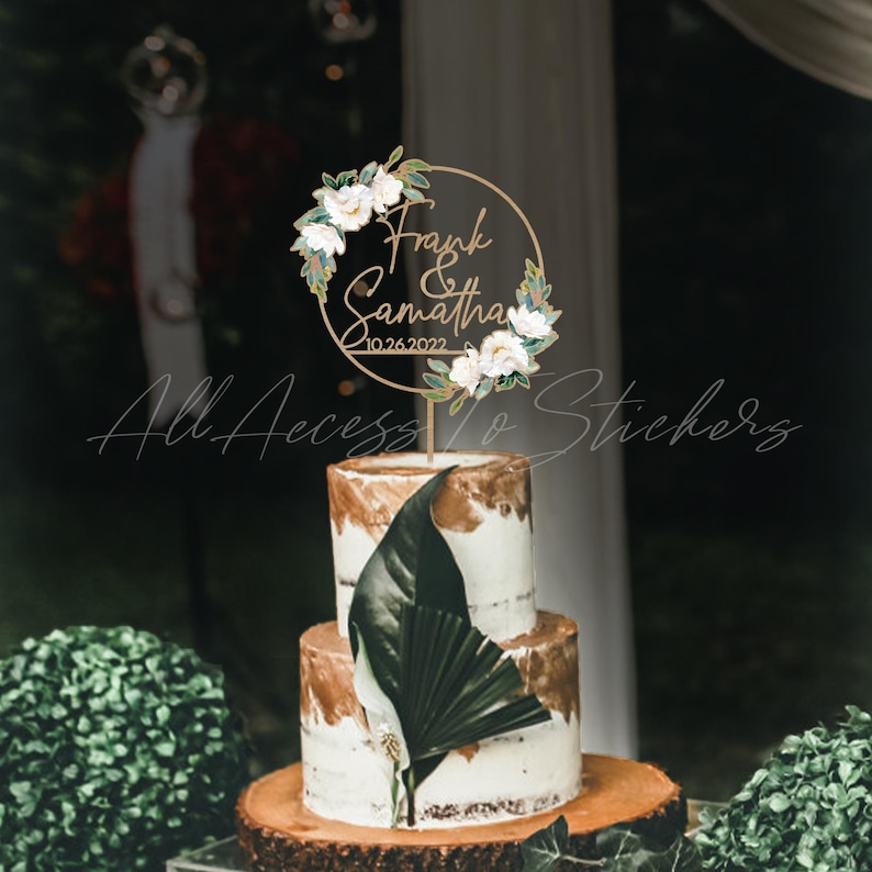 May include: A two-tiered wedding cake with a gold and white design, topped with a floral wreath cake topper. The topper has the names "Frank & Samantha" and the date "10.26.2022". A large green leaf decorates the front of the cake.