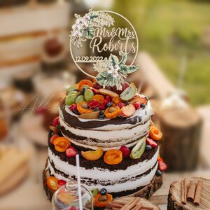 Peut inclure: Un g&acirc;teau &agrave; deux &eacute;tages avec du gla&ccedil;age blanc et des couches de g&acirc;teau au chocolat, garni de fruits frais et d'un cake topper en bois. Le cake topper indique "Mr & Mrs Roberts" et la date "12.09.2022", avec des accents floraux et de feuilles.
