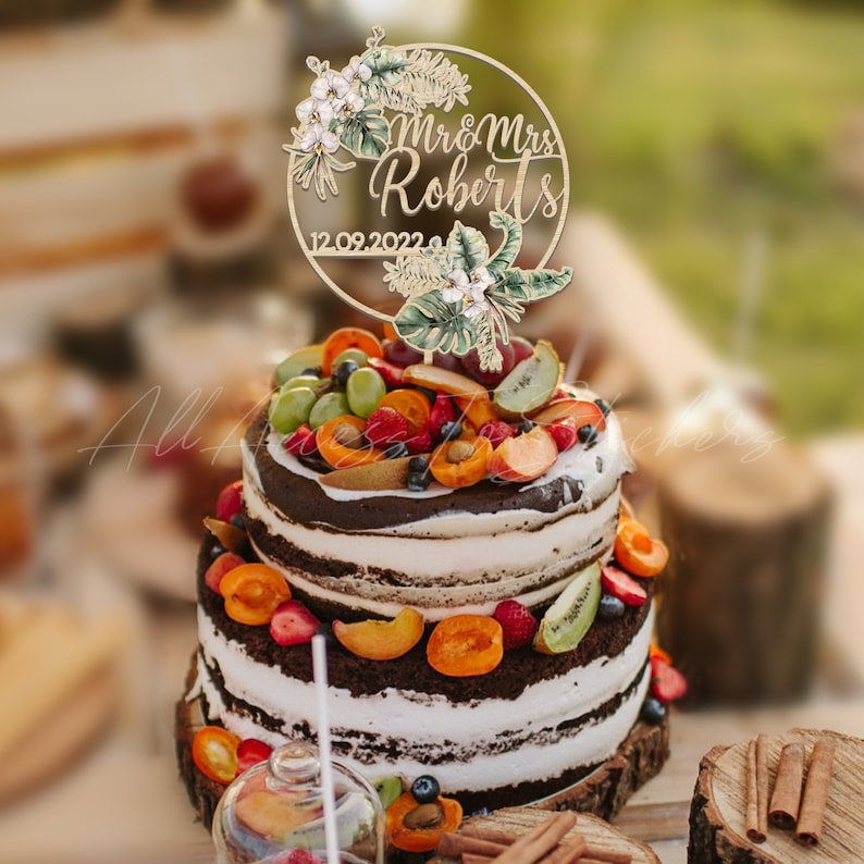 Peut inclure: Un g&acirc;teau &agrave; deux &eacute;tages avec du gla&ccedil;age blanc et des couches de g&acirc;teau au chocolat, garni de fruits frais et d'un cake topper en bois. Le cake topper indique "Mr & Mrs Roberts" et la date "12.09.2022", avec des accents floraux et de feuilles.