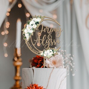 May include: A circular wooden cake topper featuring the names Jeremy and Alyssa in script lettering, the date 07.26.2023, and decorative clusters of white roses and green leaves arranged along the top and bottom edges of the circular frame.