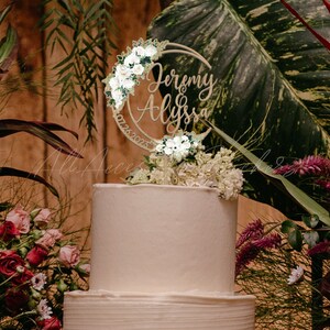 May include: A circular wooden cake topper featuring the names Jeremy and Alyssa in cursive script, the date 07.26.2023, and decorative clusters of white roses and green leaves arranged along the crescent-shaped frame atop a white tiered cake.