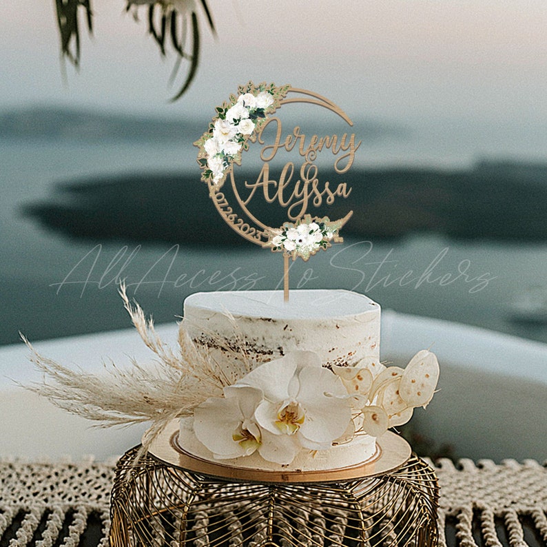 May include: A wooden cake topper with a crescent moon design and white flowers. The topper has the names 'Jeremy & Alyssa' and the date '07.28.2023'.