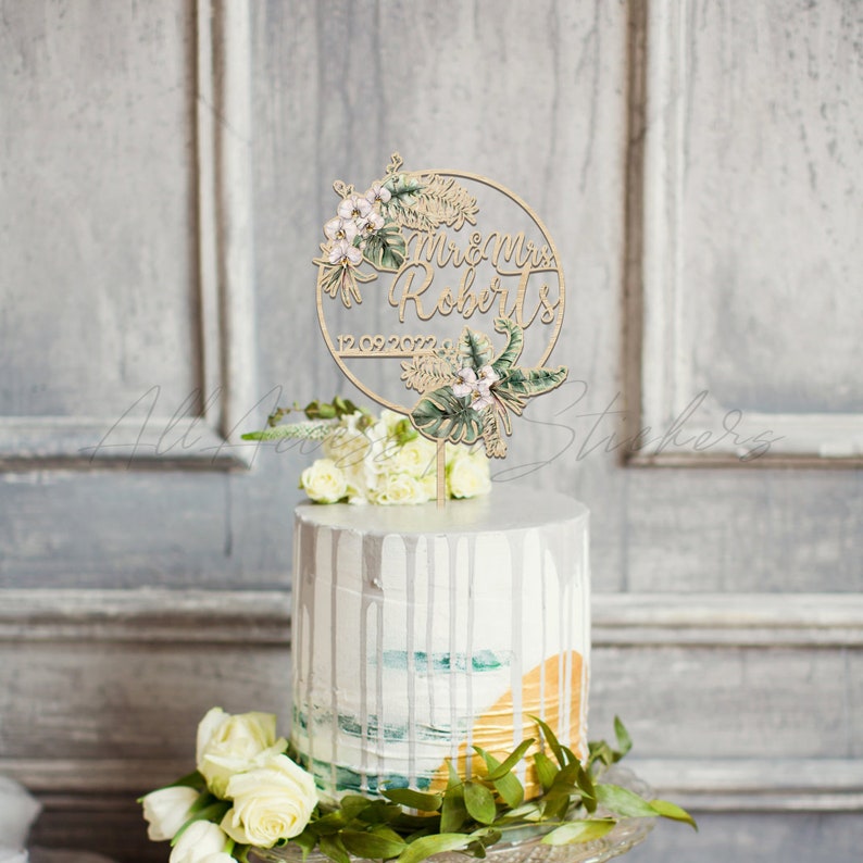 Peut inclure: Un cake topper en bois avec les mots "Mr & Mrs Roberts" et la date "12.09.2022" dans un design circulaire, d&eacute;cor&eacute; d'&eacute;l&eacute;ments floraux et de feuilles. Le topper est pos&eacute; sur un g&acirc;teau blanc avec des coulures d&eacute;coratives et des arrangements floraux.