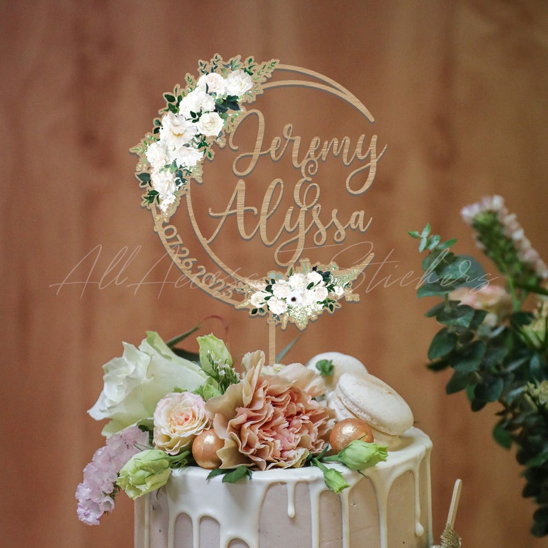 May include: A circular wooden cake topper featuring the names Jeremy and Alyssa in a script font, with the date 07.26.2025 engraved along the side. The topper is decorated with white floral arrangements on the top left and bottom right.