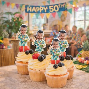 May include: Four cupcakes topped with white frosting, fresh strawberries, blueberries, and paper cutouts of a man wearing a floral shirt and holding a coconut. The cutouts feature a smiling face with a beard and a thumbs-up gesture.