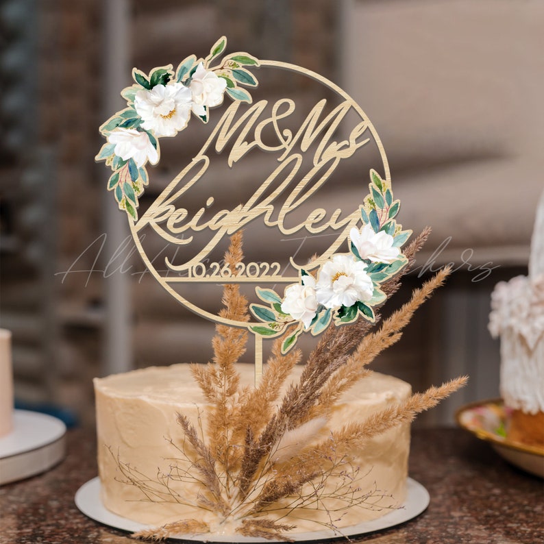 May include: A wedding cake topped with a wooden cake topper. The topper features the names "M & Mrs Keighley" and the date "10.26.2022" surrounded by a floral wreath with white flowers and green leaves. The cake is decorated with dried grasses.