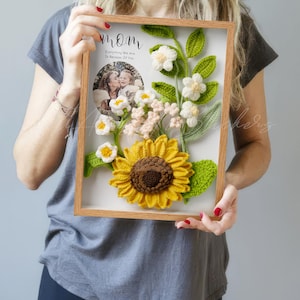 May include: A framed artwork with a crocheted sunflower and floral design. The frame includes a photo and the text "Mom Everything We Are Is Because Of You". The artwork is in a wooden frame, ideal for a gift.