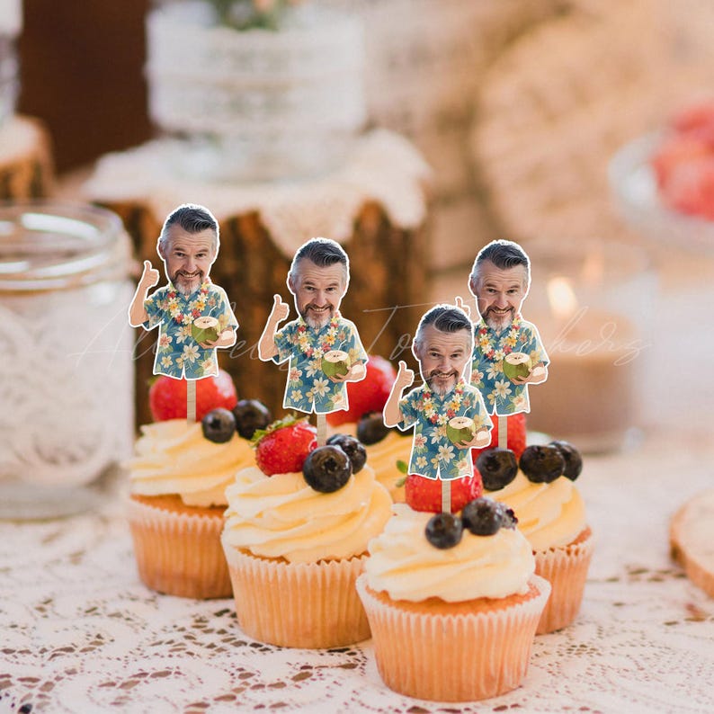 May include: Four cupcakes topped with custom paper cutouts featuring a man with a beard wearing a blue and white floral shirt, holding a green coconut. Each cupcake is decorated with fresh strawberries and blueberries on white frosting.