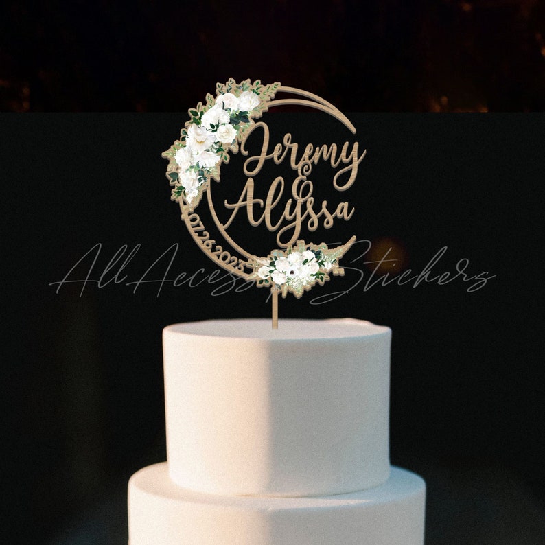 May include: A circular wooden cake topper featuring the names Jeremy and Alyssa in script font, the date 07.26.2023, and white floral accents with green leaves arranged along the crescent-shaped frame, positioned atop a plain white tiered cake.