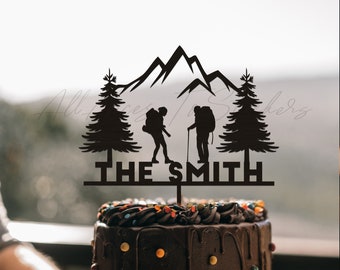 May include: A black cake topper featuring a mountain range, two trees, and silhouettes of hikers. The text "THE SMITH" is displayed below the hikers. The topper is placed on a chocolate cake with colorful sprinkles.