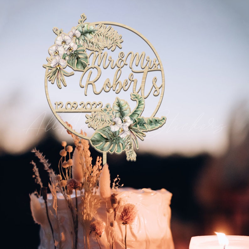 Peut inclure: Un cake topper en bois avec l'inscription "Mr & Mrs Roberts" et la date "12.09.2022" est d&eacute;cor&eacute; d'&eacute;l&eacute;ments floraux &agrave; l'aquarelle. Le cake topper est pos&eacute; sur un g&acirc;teau glac&eacute; blanc, avec des fleurs s&eacute;ch&eacute;es et une bougie en arri&egrave;re-plan.