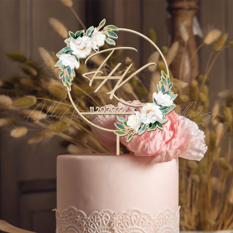 May include: A wooden wedding cake topper with a circular design. The topper features the initials "F H" and the date "11.20.2022". It is adorned with white flowers and green leaves, and is placed on top of a pink cake.