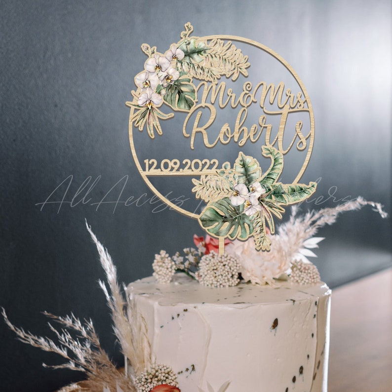 Peut inclure: Un cake topper en bois avec l'inscription "Mr & Mrs Roberts" et la date "12.09.2022" dans un cercle. Il est orn&eacute; de motifs floraux et de feuilles &agrave; l'aquarelle, notamment des orchid&eacute;es blanches et des feuilles de monstera vertes. Le g&acirc;teau est blanc avec des &eacute;l&eacute;ments d&eacute;coratifs.