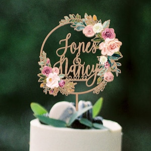 Puede incluir: Un adorno circular de madera para pastel con el texto Jones & Nancy y la fecha 20.07.22 en letra cursiva. El adorno est&aacute; decorado con un arreglo floral de flores rosas, blancas y color melocot&oacute;n con hojas verdes en el lado derecho.