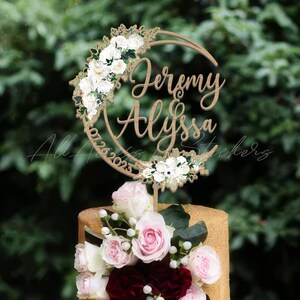 May include: A circular wooden cake topper featuring the names Jeremy and Alyssa in script font, with the date 07.26.2023 engraved along the side. The topper is decorated with white floral accents and sits atop a cake adorned with pink roses.