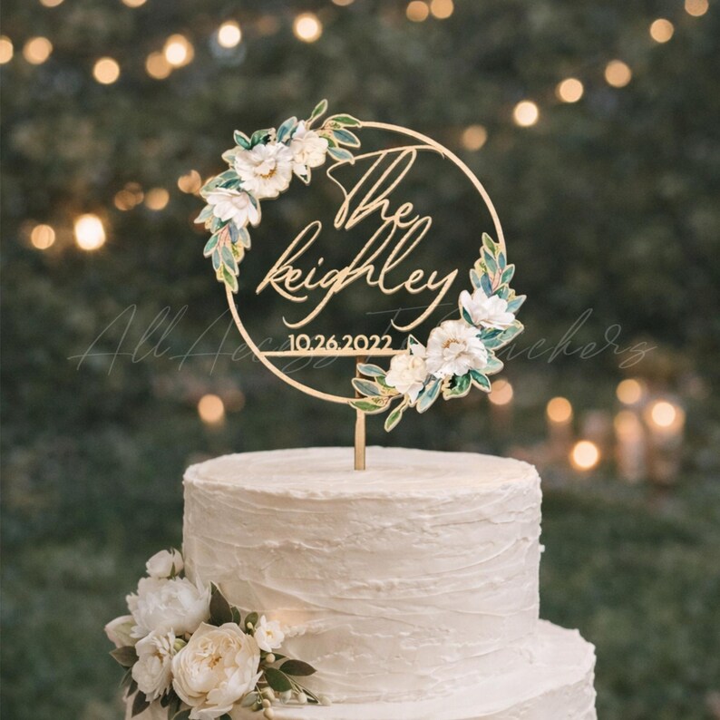 May include: A gold-colored cake topper with the words "The Keighley" and the date "10.26.2022" in a circular design. The circle is adorned with white flowers and green leaves. The topper is placed on a white tiered cake.
