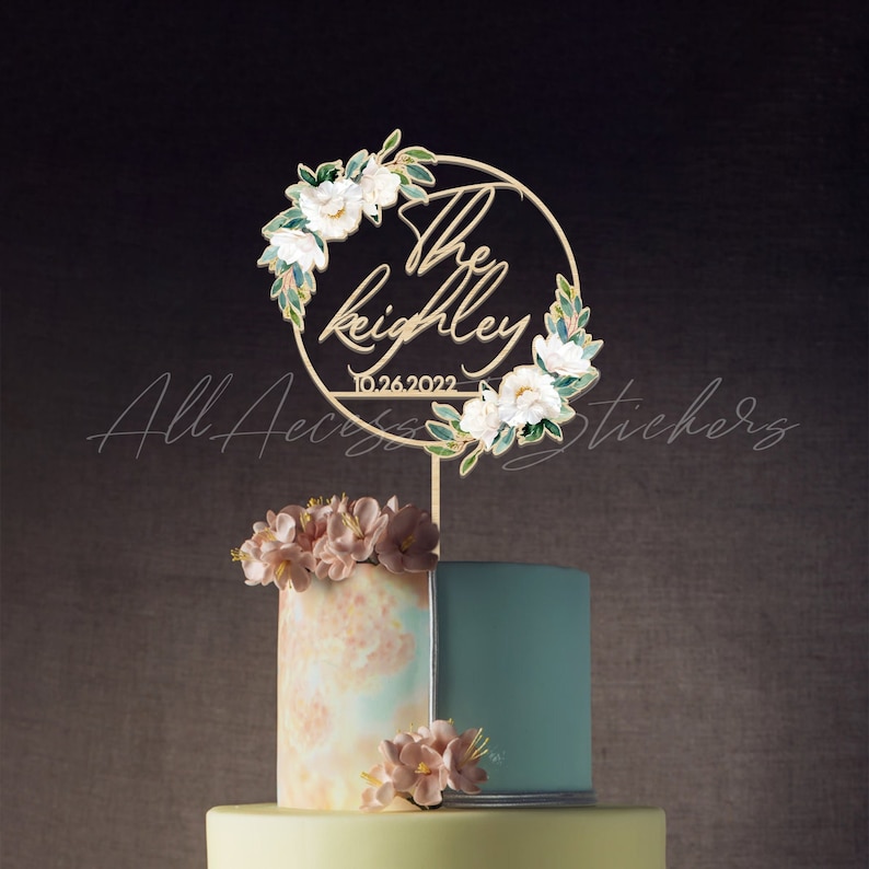 May include: A gold-colored cake topper with the names "The Keighley" and the date "10.26.2022" in a circular design, adorned with white flowers and green leaves. The topper is placed on a multi-tiered cake with floral decorations.