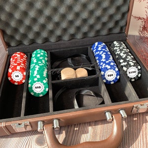 May include: A brown carrying case with a foam-lined lid containing stacks of poker chips in red, green, blue, and black. The chips are labeled with $5, $25, $50, and $100 denominations. Two wooden dice are placed in the center compartment.