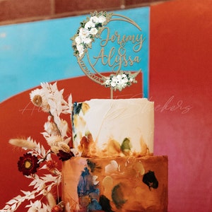 May include: A circular wooden cake topper featuring the names Jeremy and Alyssa in cursive script, the date 07/26/2023, and floral accents with white roses and greenery. The topper is positioned on a two-tiered cake with abstract paint-like patterns.