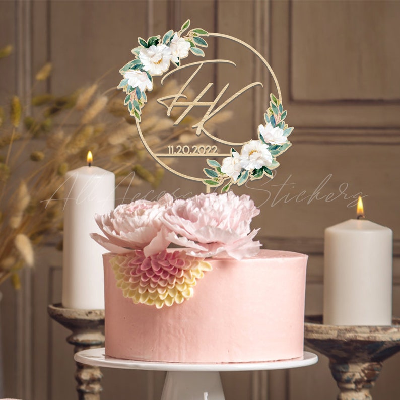 May include: A pink frosted cake with a floral decoration and a wooden cake topper. The topper features initials within a floral wreath and the date 11.20.2022. Two white candles are on either side of the cake.