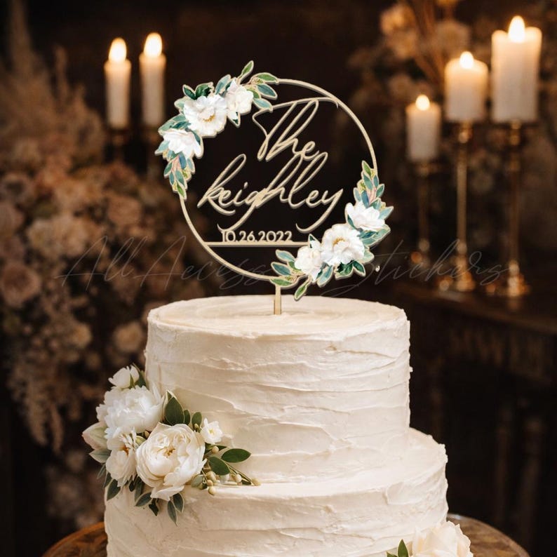 May include: A two-tiered white wedding cake with a floral arrangement and a gold cake topper. The topper is a circular design with the names "The Keighley" and the date "10.26.2022" in script, surrounded by white flowers and green leaves.
