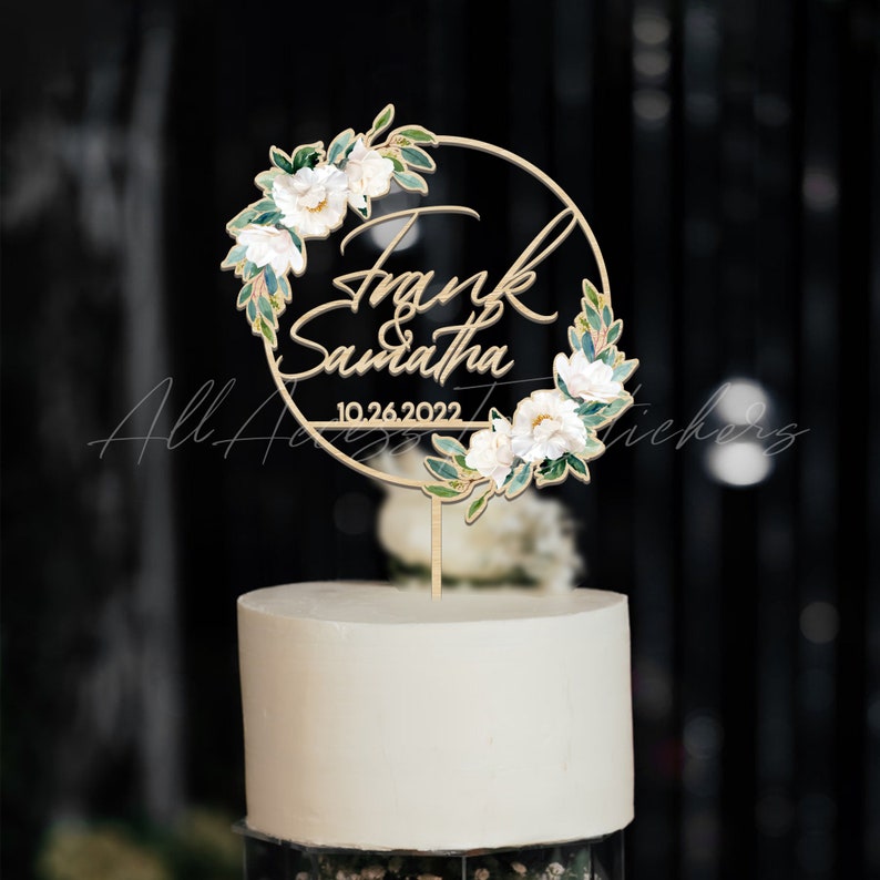 May include: A wedding cake topped with a wooden cake topper. The topper is a gold-colored circle with the names "Frank & Samantha" and the date "10.26.2022" in script. The circle is decorated with white flowers and green leaves.
