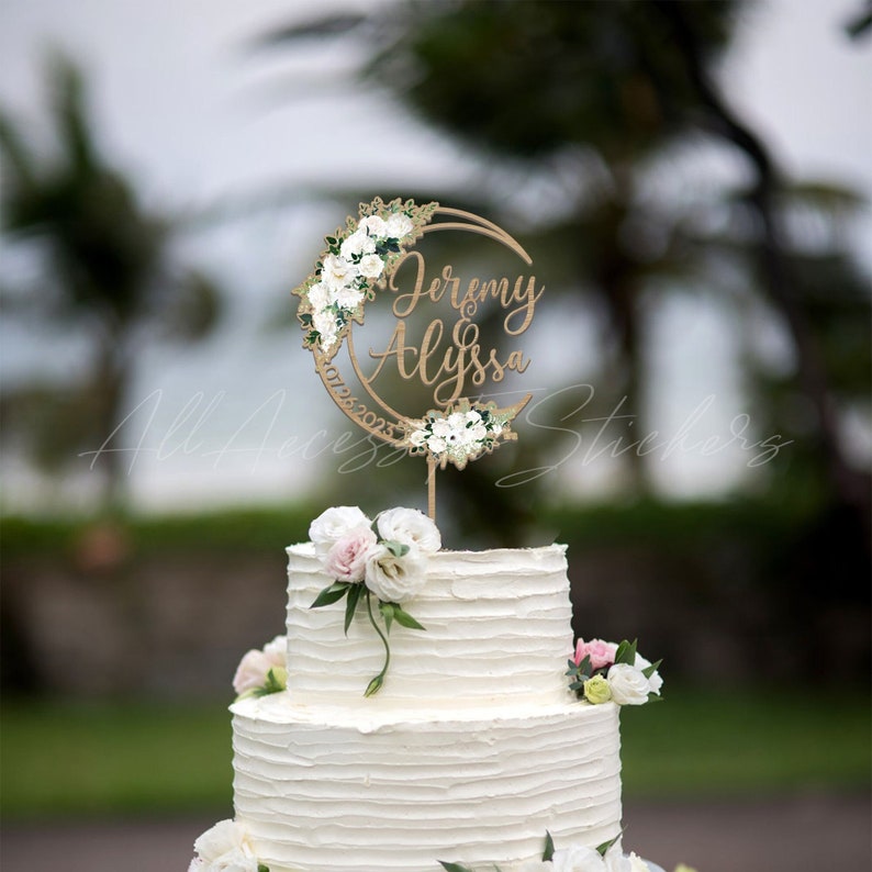 May include: A two-tier white frosted cake topped with a circular wooden cake topper featuring the names Jeremy and Alyssa, the date 10/25/2023, and decorative white floral accents. The cake is adorned with fresh white and light pink flowers.