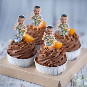 May include: Four cupcakes with chocolate frosting, topped with small brown candy spheres, orange slices, and paper cutouts of a person wearing a floral shirt, giving a thumbs up, and holding a coconut. The cupcakes are in white paper liners.
