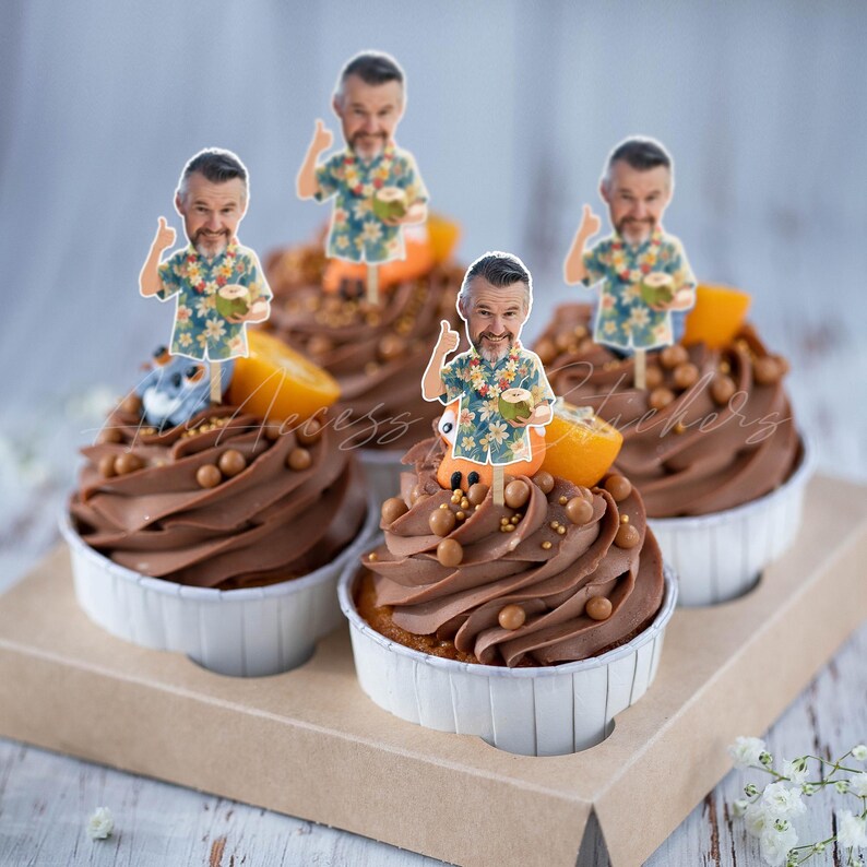 May include: Four cupcakes with chocolate frosting, topped with small brown candy spheres, orange slices, and paper cutouts of a person wearing a floral shirt, giving a thumbs up, and holding a coconut. The cupcakes are in white paper liners.