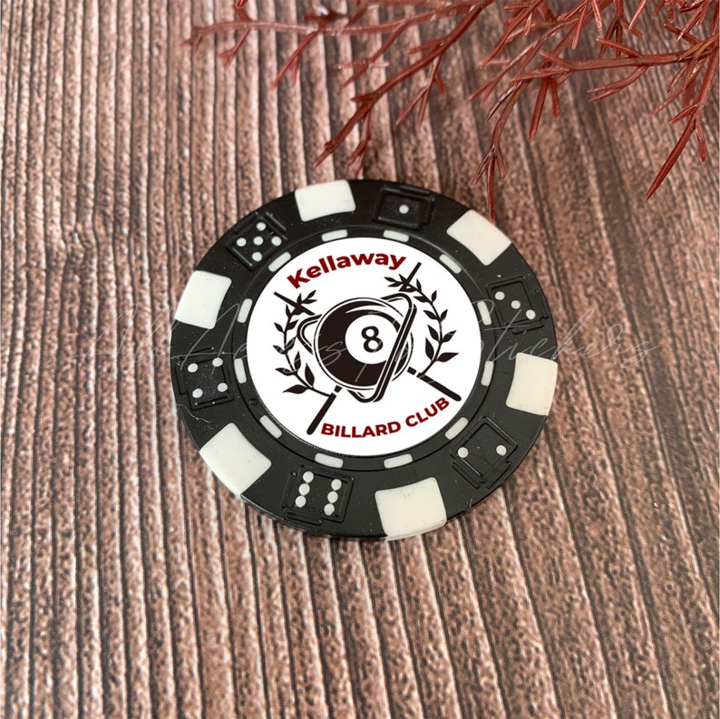 May include: A round black and white poker chip featuring a central graphic of an 8-ball surrounded by laurel branches and the text Kellaway BILLARD CLUB. The outer edge of the chip is decorated with small dice patterns in alternating black and white.