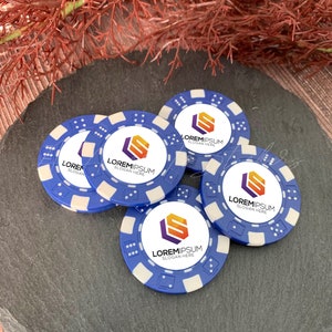 May include: Four circular blue poker chips with white edges and dice-patterned accents. Each chip features a central white circular inlay printed with a hexagonal orange and purple logo, the text LOREMIPSUM, and the smaller text SLOGAN HERE.