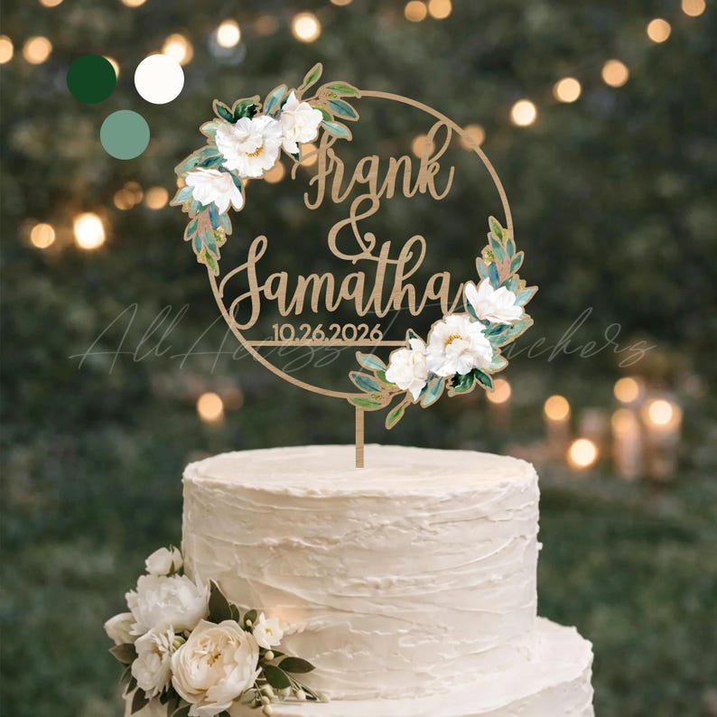 May include: A wooden cake topper with the names "Frank & Samantha" and the date "10.26.2026" in elegant script. The topper is encircled by a floral wreath with white flowers and green leaves, placed atop a tiered white cake.