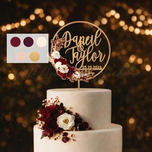 May include: A circular wooden cake topper featuring the names "Daneil & Taylor" and the date "05.25.2024" in script lettering. The topper is decorated with a cluster of artificial burgundy and white flowers and gold-toned foliage.