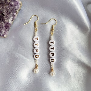 May include: Dangle earrings with gold-tone hooks. Each earring has white letter beads spelling "TTPD" in gold, with a small pearl at the base. The earrings are presented on a light grey, silky fabric.