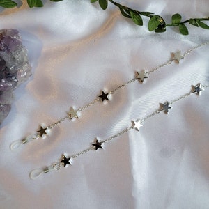 May include: Two silver eyeglass chains with star-shaped charms. The chains are displayed on a white satin surface, with a sprig of green leaves in the background. A purple crystal is also visible.