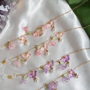 May include: Gold-coloured eyeglass chains adorned with floral and butterfly motifs. The chains feature pink, purple, and clear flowers, butterflies, and faux pearls. Clear rubber loops are attached for securing to glasses.