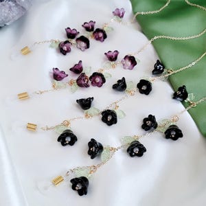 以下が含まれることがあります： 黒と紫の花のアクセントが付いた、金色のメガネチェーン。チェーンは金色の色で、小さな緑の葉のディテールがあります。メガネホルダーは透明で、金色のコイルが付いています。背景は白と緑です。