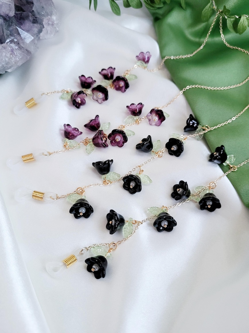 以下が含まれることがあります： 黒と紫の花のアクセントが付いた、金色のメガネチェーン。チェーンは金色の色で、小さな緑の葉のディテールがあります。メガネホルダーは透明で、金色のコイルが付いています。背景は白と緑です。