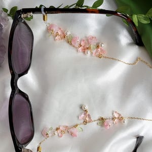 May include: Eyeglass chain with a floral design. The chain features pink flowers, green leaves, and faux pearls. The chain is attached to a pair of tortoiseshell eyeglasses.