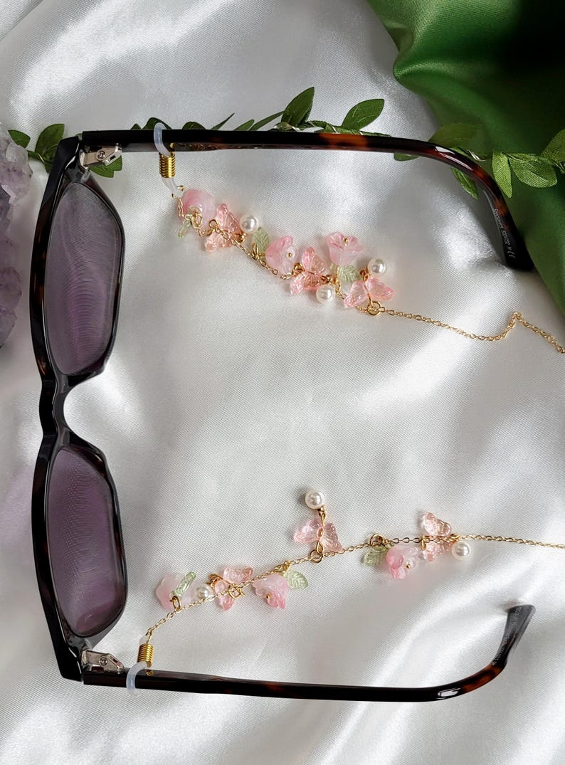 May include: Eyeglass chain with a floral design. The chain features pink flowers, green leaves, and faux pearls. The chain is attached to a pair of tortoiseshell eyeglasses.