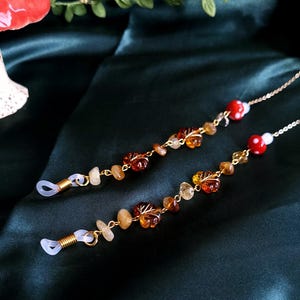 May include: Two eyeglass chains with gold-colored metal chains and clear rubber loops. The chains are adorned with amber-colored leaf and stone beads, and red mushroom beads. The background is a dark green fabric.