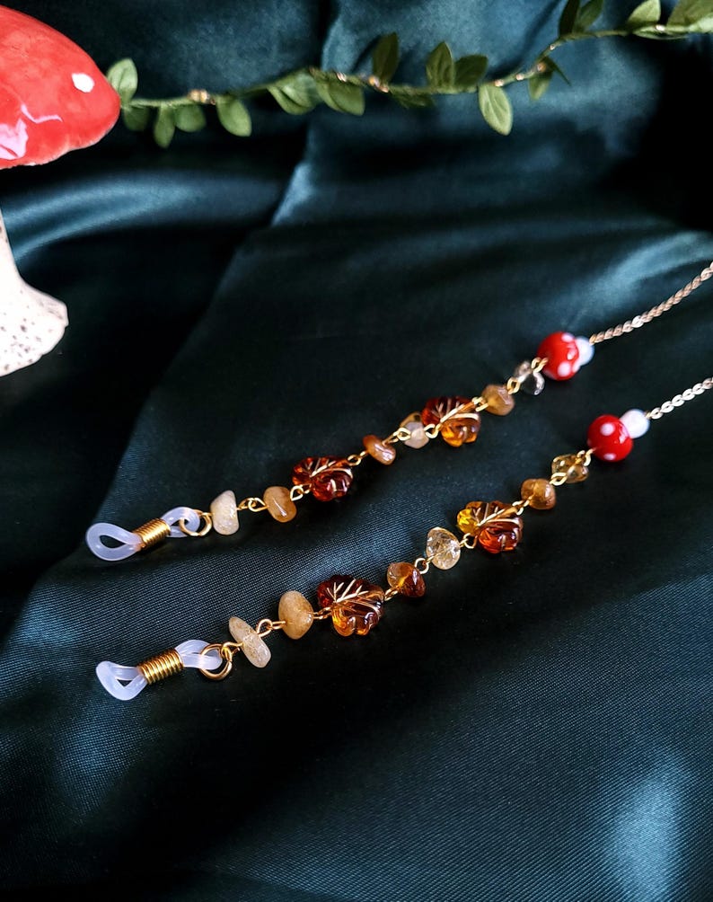 May include: Two eyeglass chains with gold-colored metal chains and clear rubber loops. The chains are adorned with amber-colored leaf and stone beads, and red mushroom beads. The background is a dark green fabric.