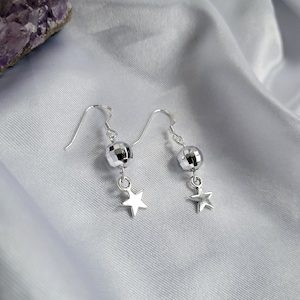 May include: A pair of silver earrings with a disco ball-shaped bead and a star charm. The earrings are hanging on a white satin surface. The earrings are a shiny silver color.
