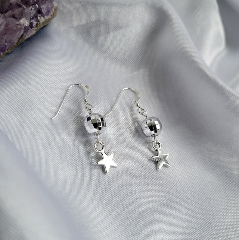 May include: A pair of silver earrings with a disco ball-shaped bead and a star charm. The earrings are hanging on a white satin surface. The earrings are a shiny silver color.