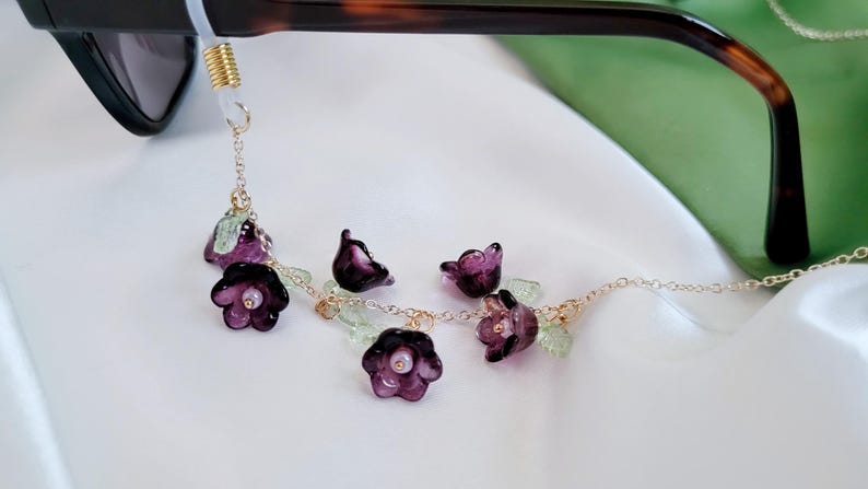 以下が含まれることがあります： 紫色の花と緑の葉のアクセントが付いた、ゴールドトーンのメガネチェーン。チェーンはサングラスに接続されています。花は半透明で、中央に小さな白いビーズが施されています。