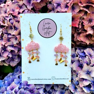May include: Pink cloud earrings with gold sunburst details, gemstone drops, and butterfly charms. The earrings are displayed on a white card with "Laura Gordon Art" and social media details. The background features pink and purple flowers.