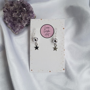 May include: Silver-toned earrings with a disco ball and star charm. The earrings hang from a silver hook and are displayed on a white card with the text "Laura Gordon Art" in a pink circle. A purple crystal is in the background.