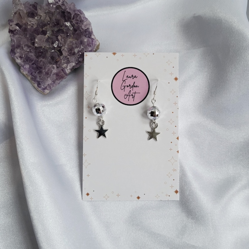 May include: Silver-toned earrings with a disco ball and star charm. The earrings hang from a silver hook and are displayed on a white card with the text "Laura Gordon Art" in a pink circle. A purple crystal is in the background.