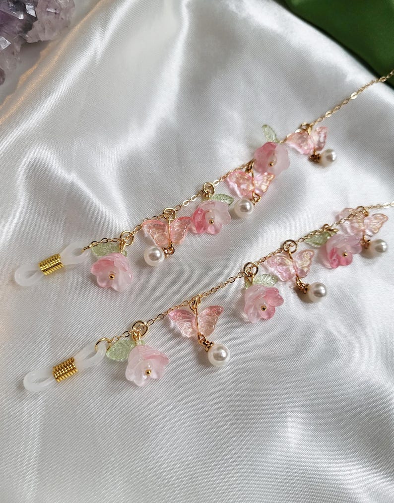 May include: Two eyeglass chains with delicate gold-coloured chains. Each chain features pink flowers, butterflies, green leaves, and faux pearls. The chains are designed to hold eyeglasses and add a decorative touch.